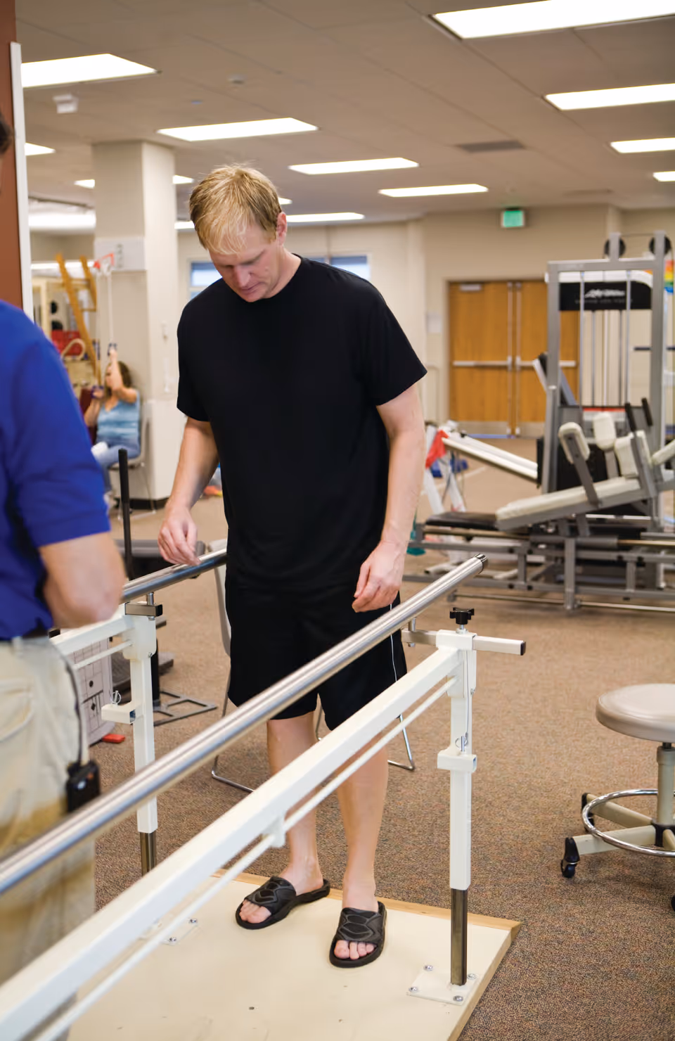 A man in a black t-shirt and shorts is using parallel bars for walking exercise in a rehabilitation or physical therapy room. Another person, partially visible, is standing nearby. The room contains exercise equipment and has a carpeted floor with fluorescent ceiling lights.