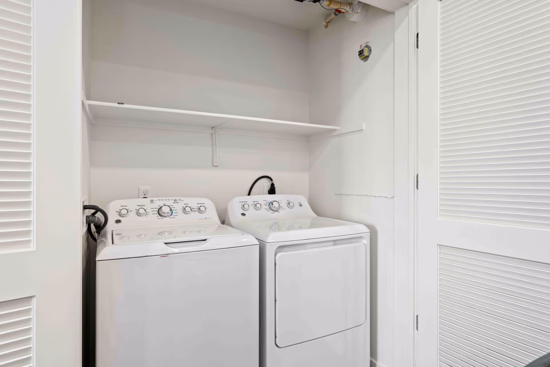A small laundry area with a white washing machine and dryer side by side, white walls, a white shelf above the machines, and white louvered doors partially open on either side.