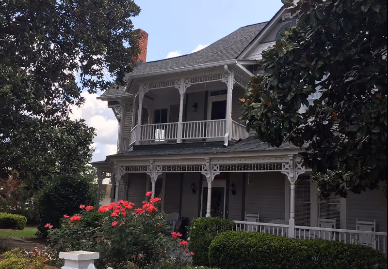 A two-story house with a large porch and balcony featuring white railings and decorative trim. The house is surrounded by green bushes and blooming red flowers, with a clear blue sky in the background.
