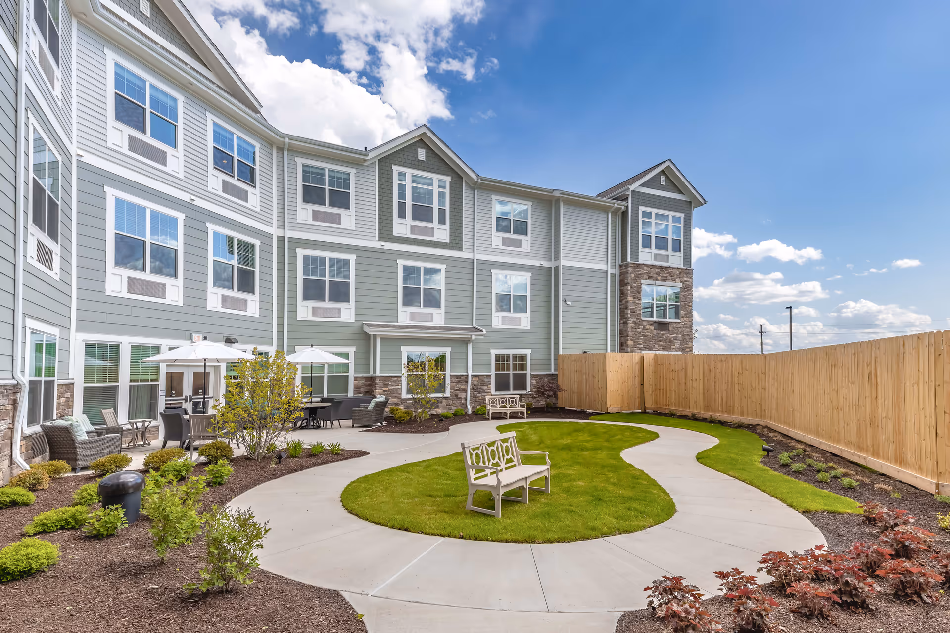 Courtyard with a curved concrete walkway, benches, patio seating and umbrellas beside a three-story senior living building under a blue sky.