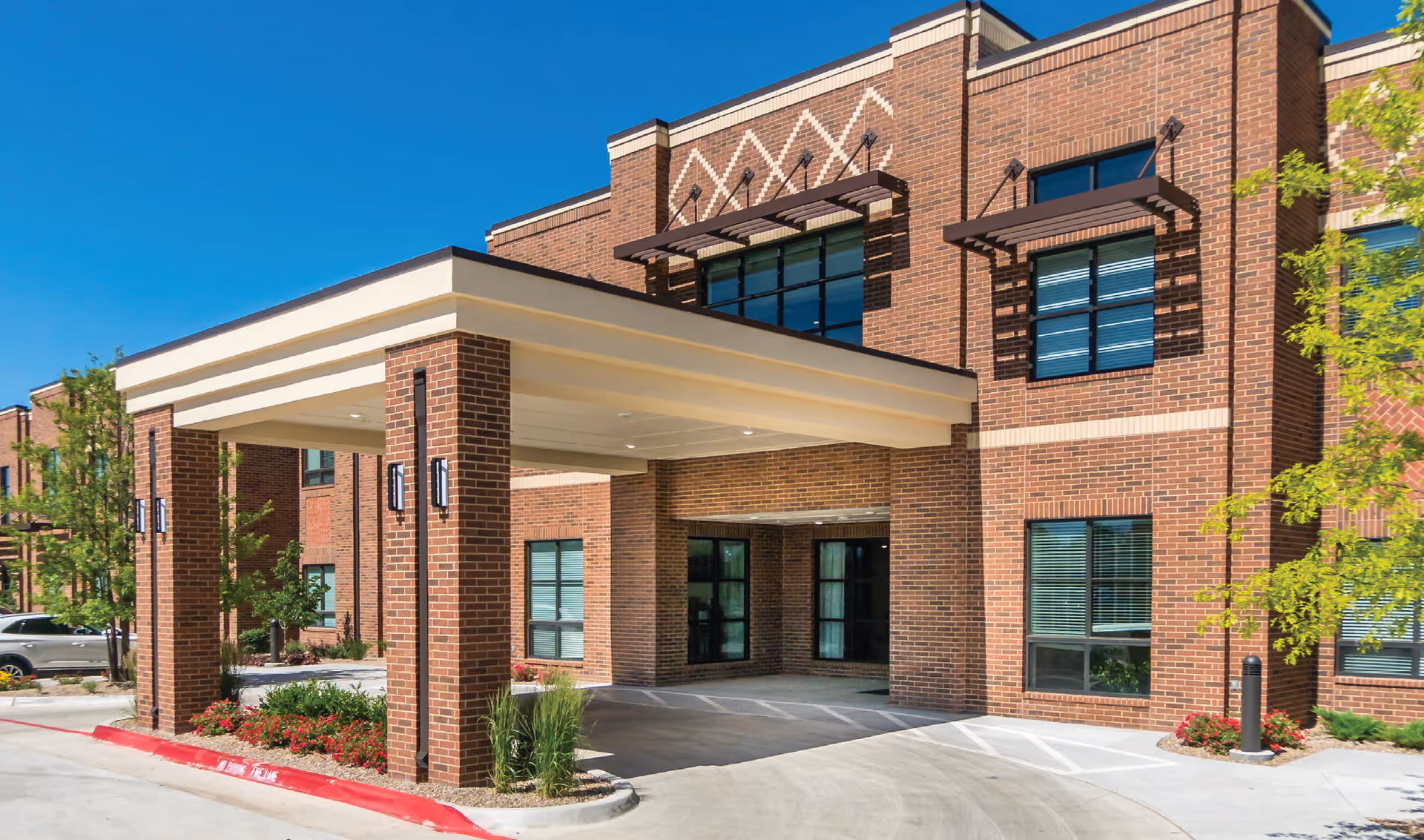Brick building front with a covered drive-up entrance, windows, landscaping, and a clear blue sky.