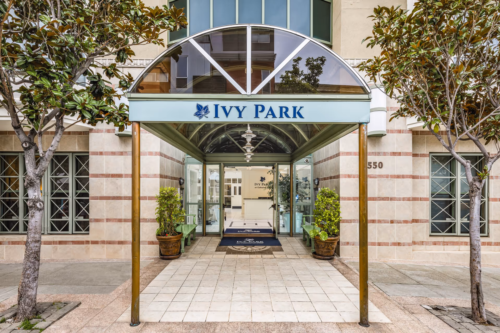 Entrance to Ivy Park at Cathedral Hill facility with a covered walkway supported by metal poles, potted plants on either side, and glass doors leading inside. The building exterior features tiled walls and windows with decorative grilles. Two trees flank the entrance.