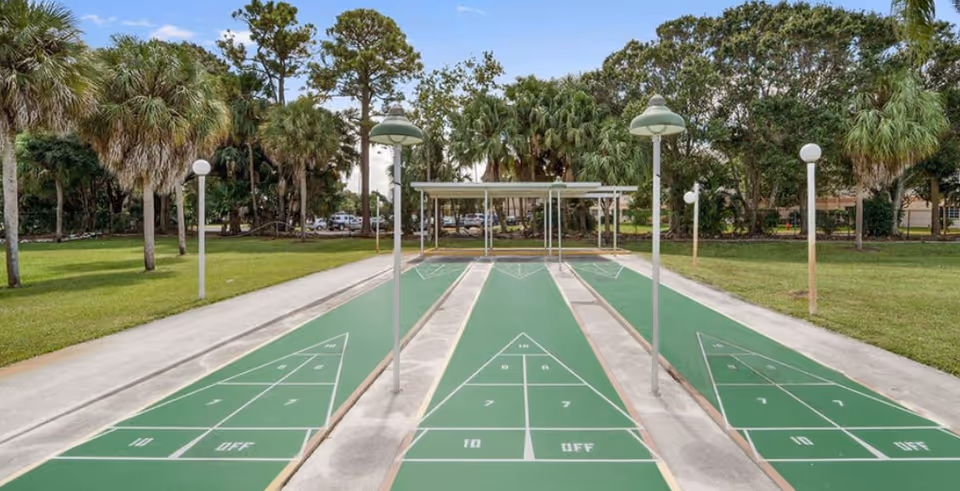 Three shuffleboard courts with green playing surfaces and white markings, surrounded by a grassy area with palm trees and other trees under a blue sky with some clouds.