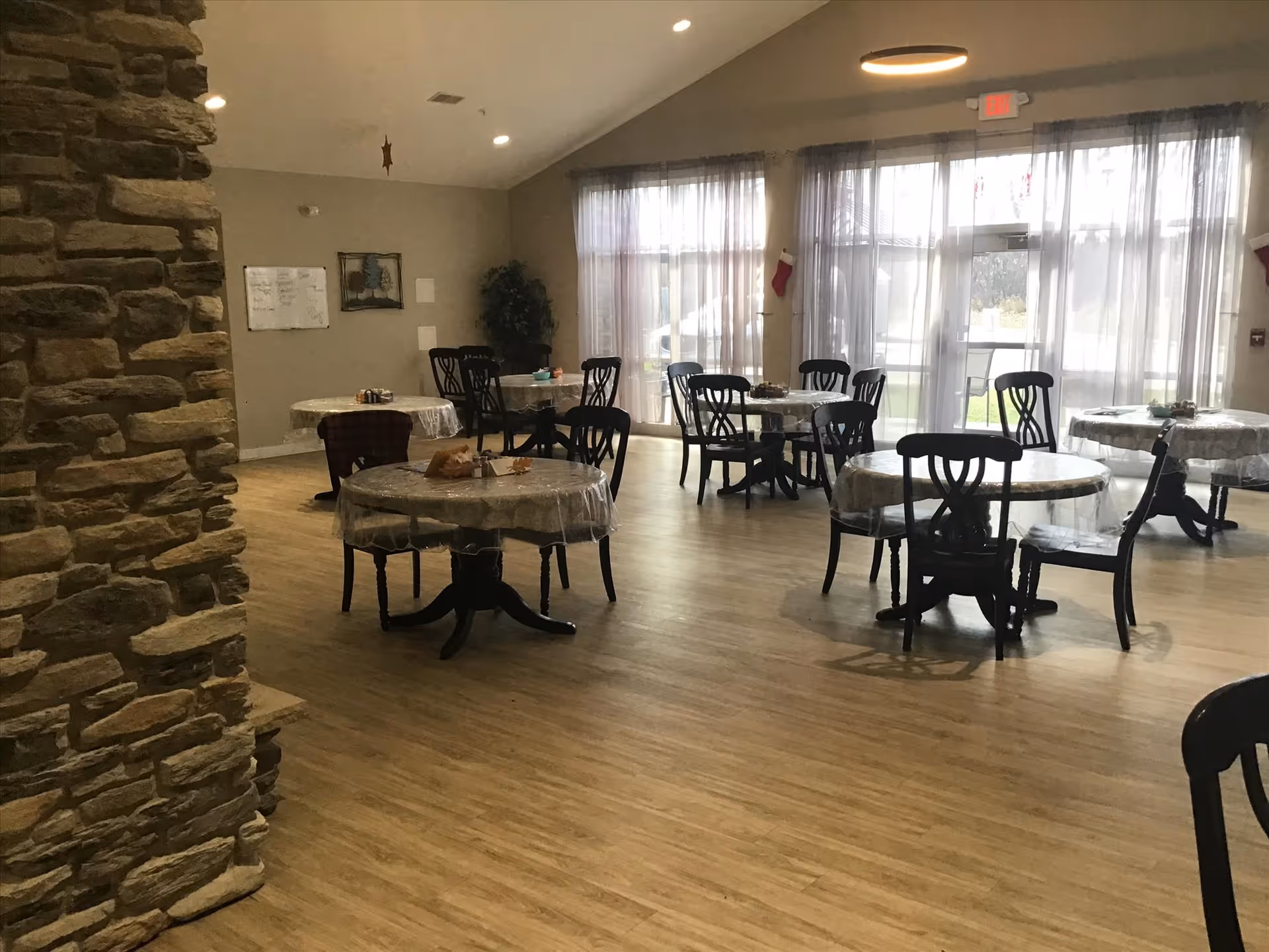 Interior view of a dining room with several round tables covered with plastic tablecloths and surrounded by black chairs. The room has wooden flooring, large windows with sheer curtains allowing natural light, and a stone wall on the left side. There are some decorations on the tables and a plant in the corner.