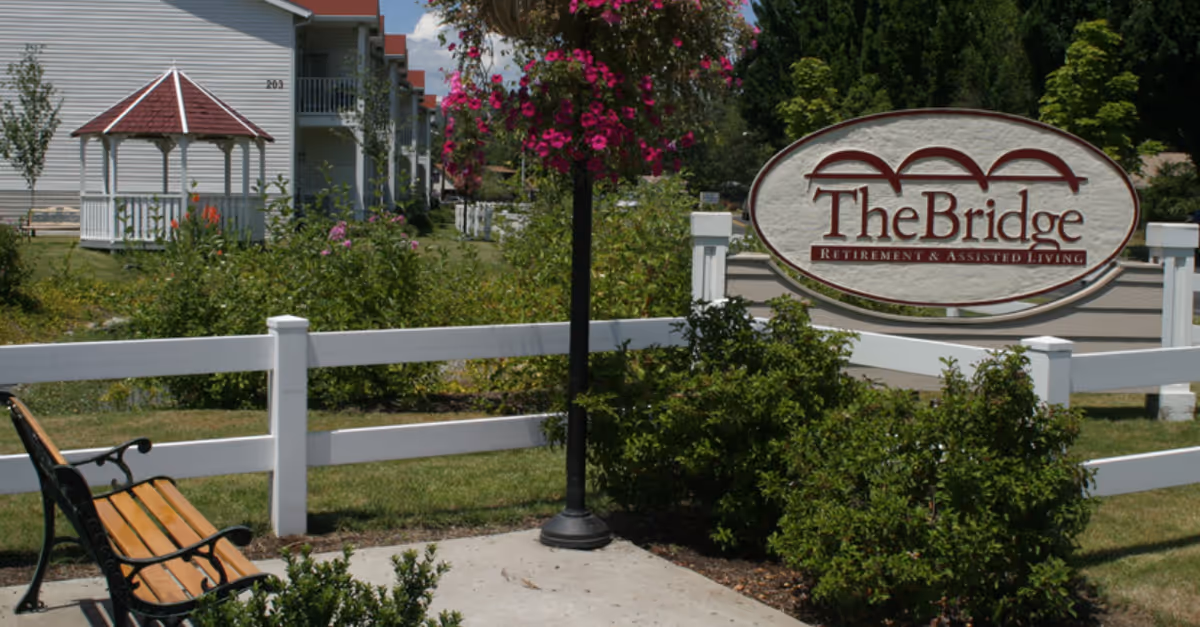 Outdoor area of The Bridge Retirement & Assisted Living facility featuring a wooden bench with black metal armrests, a white fence, green bushes, a hanging basket with pink flowers, a lamppost, and a white gazebo in the background near residential buildings.