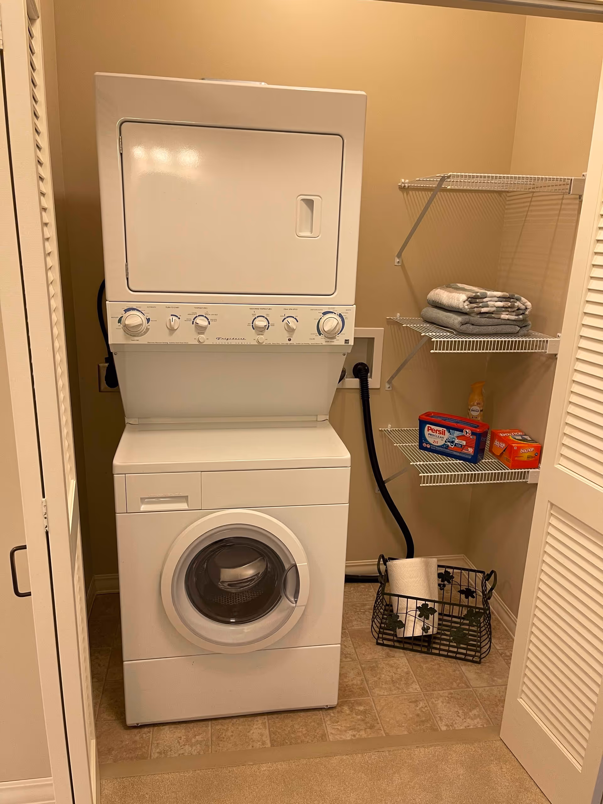 Stacked washer and dryer in a small laundry closet with wire shelves holding towels and detergent.