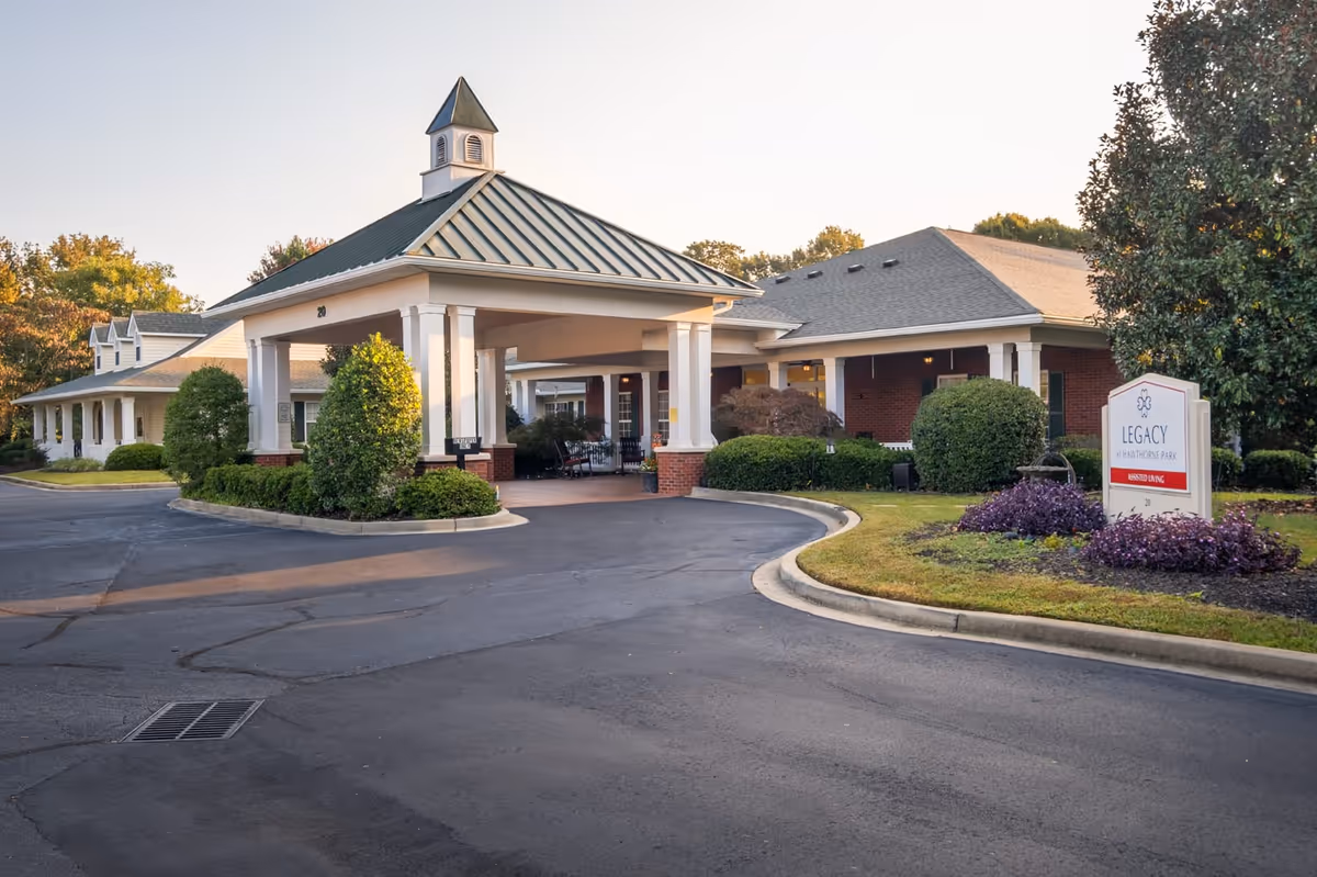 Front entrance with a covered porte-cochere, circular driveway, landscaping, and a sign for the senior living facility.