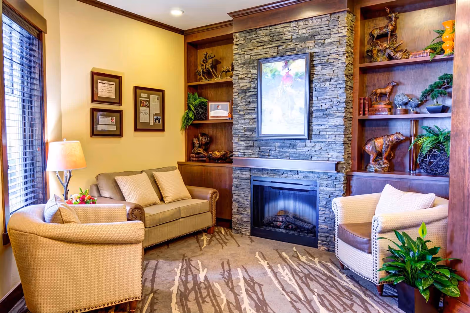 Cozy living room area with a stone fireplace in the center, flanked by built-in wooden shelves decorated with animal figurines, books, and plants. The room has a beige loveseat with two cushions, two beige armchairs, a floor lamp, framed pictures on the wall, and a large window with wooden blinds letting in natural light.