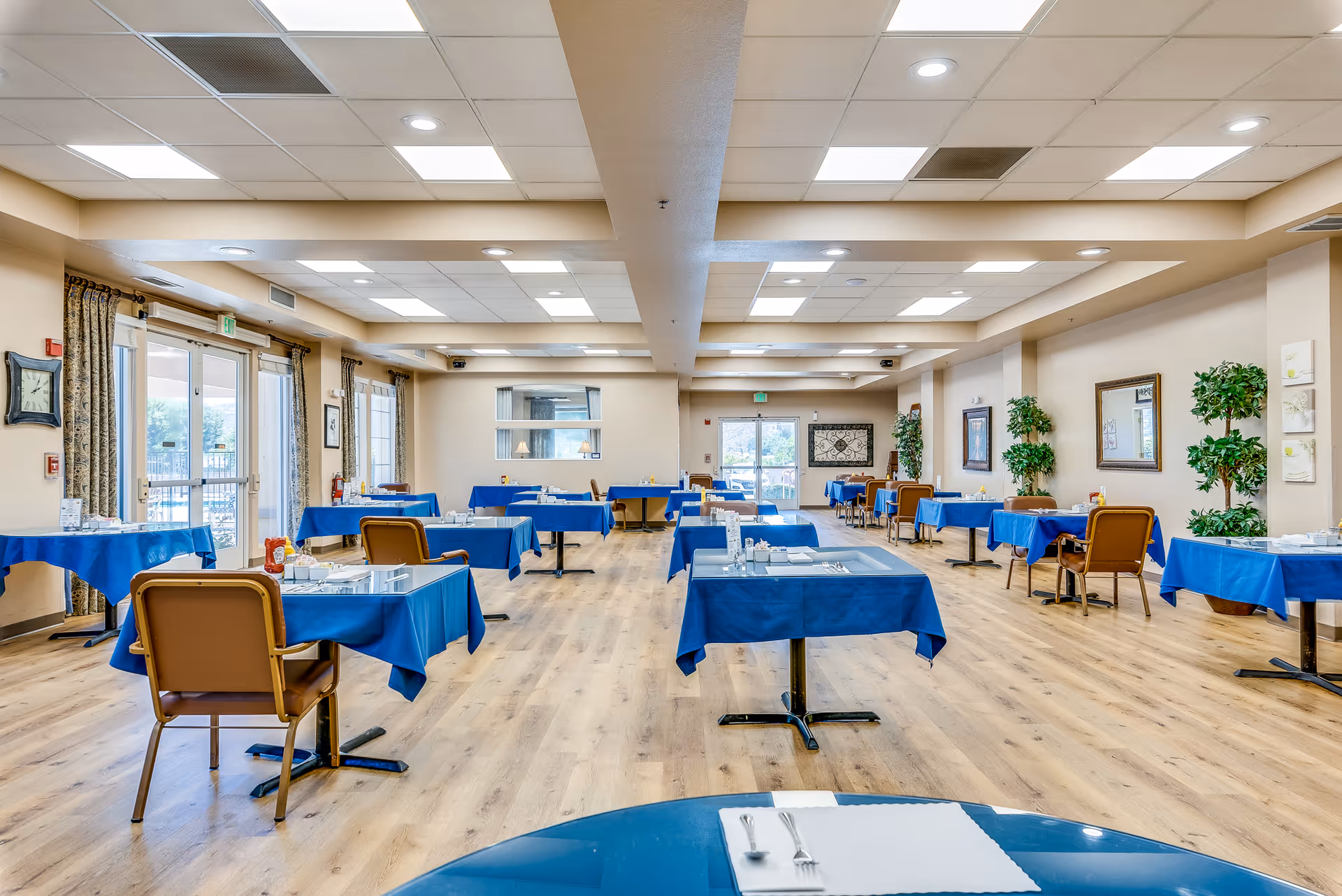 Spacious dining room with multiple tables covered in blue tablecloths and chairs arranged in a well-lit senior living facility.
