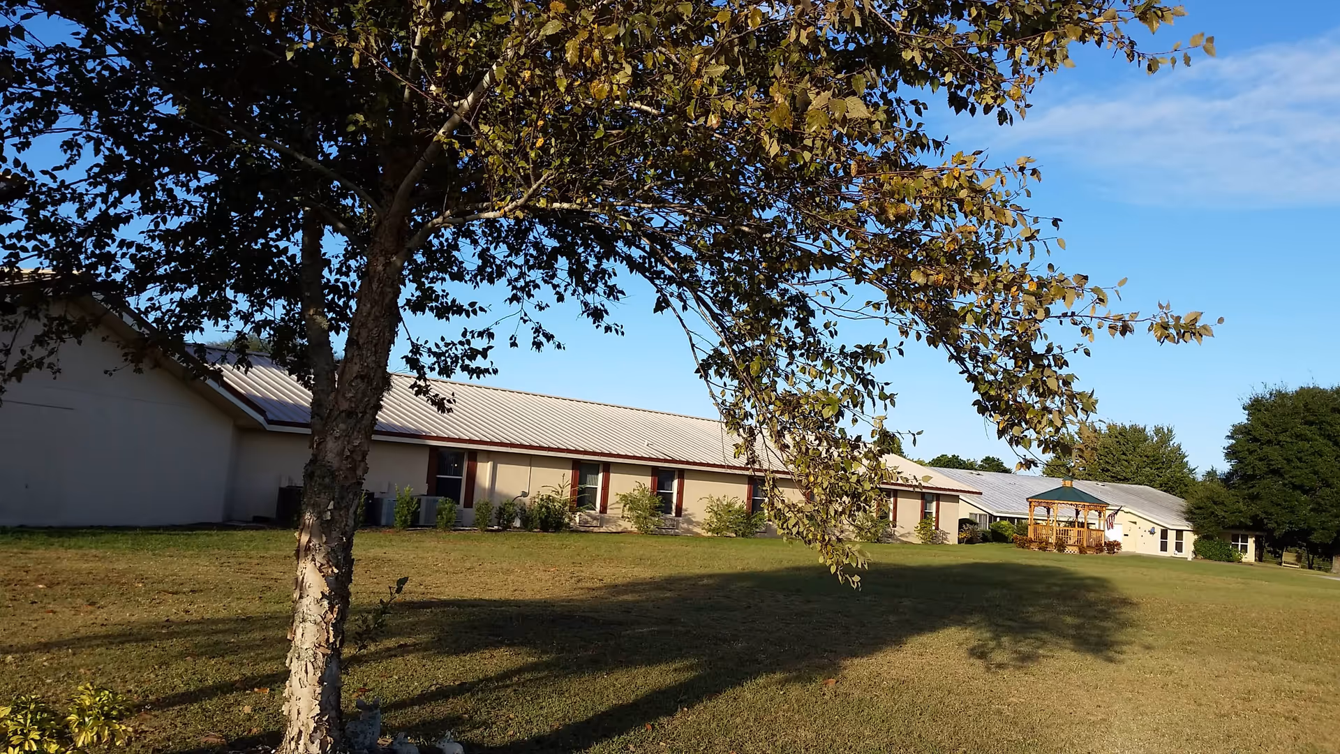 A single-story building with a light-colored roof and beige walls, partially shaded by a tree in the foreground. There is a wooden gazebo on the right side of the building, set on a large grassy lawn under a clear blue sky.