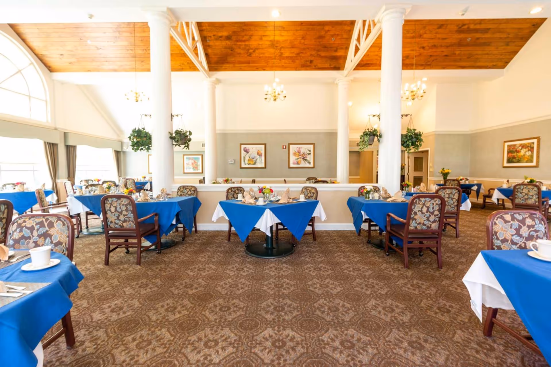 Bright dining room with tables draped in blue tablecloths, floral-upholstered chairs, tall white columns, hanging plants, and a wooden ceiling.