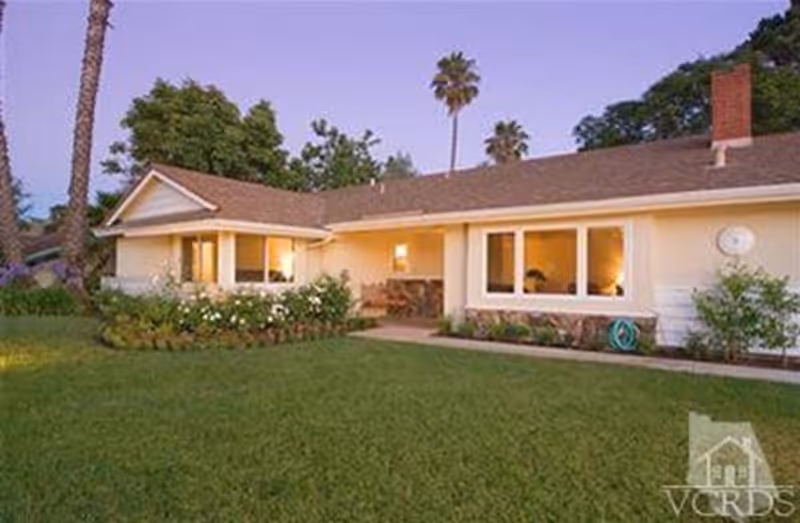 Single-story house with a well-maintained lawn and garden in front, featuring palm trees and bushes. The house has a beige exterior with large windows and a chimney, illuminated by warm indoor lighting at dusk.
