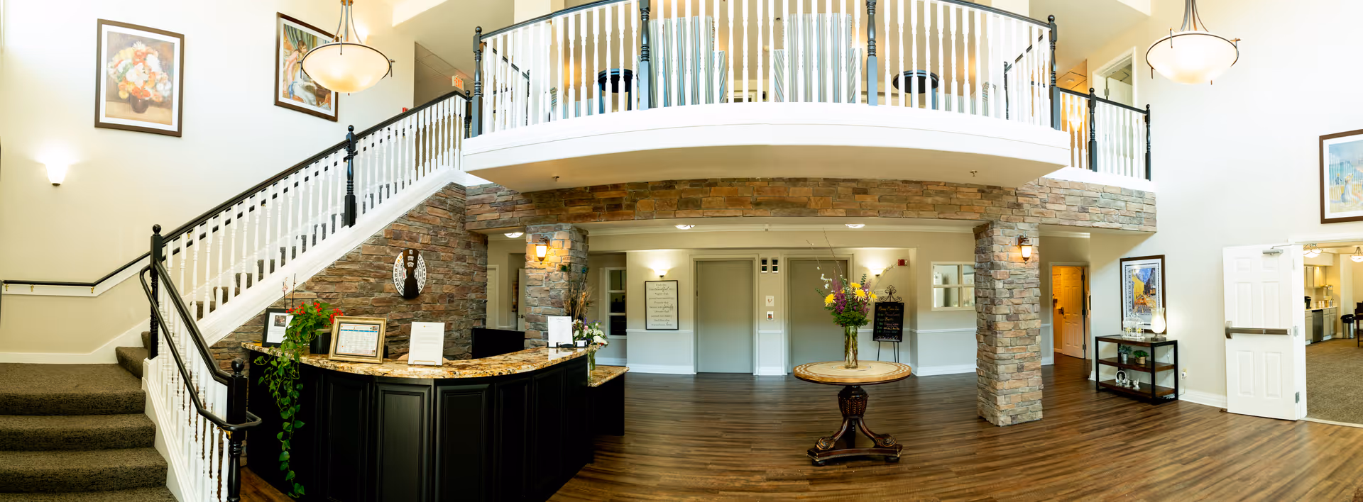 Spacious two-story senior living lobby with a reception desk, central round table with flowers, staircase and upper balcony.