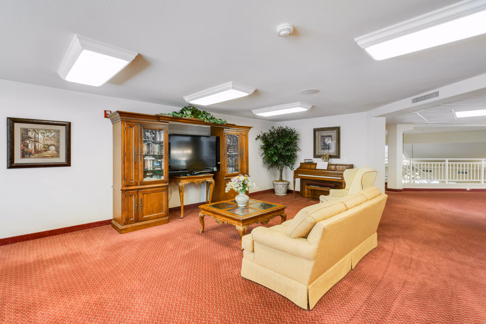 A cozy living room area with a yellow sofa and armchair facing a wooden entertainment center with a flat-screen TV. There is a wooden coffee table with a vase of flowers in front of the sofa. A piano with a bench is placed against the wall, next to a large potted plant. The room has red carpet flooring, white walls, and ceiling lights.
