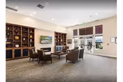 Spacious assisted-living common room with chairs and a coffee table facing a wall-mounted TV, built-in shelving with decor, and glass exit doors.