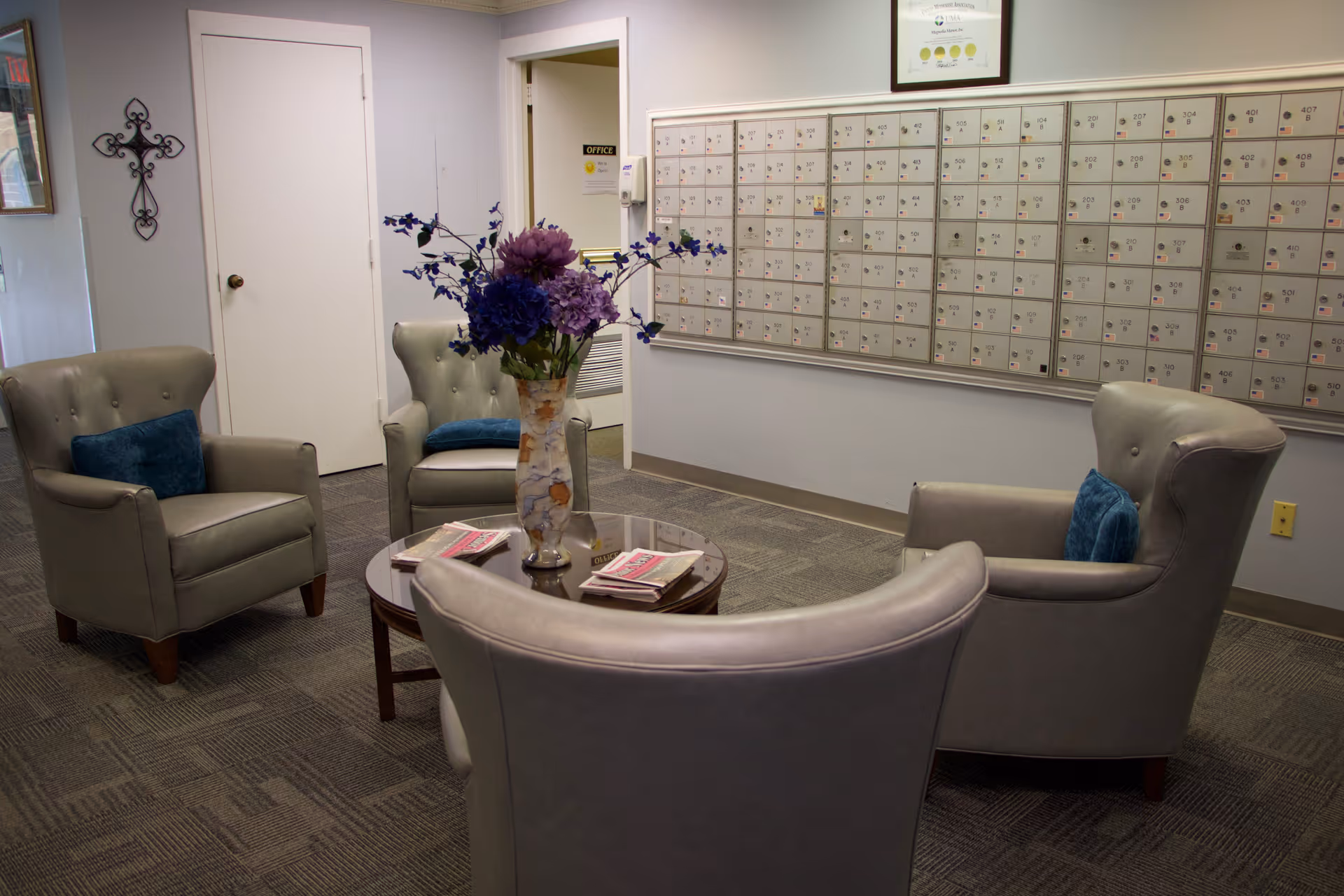 A small seating area with four gray armchairs arranged around a round glass-top table with a vase of purple and blue flowers. Behind the seating area is a wall with multiple mailboxes. There is a door labeled 'Office' and a decorative cross on the wall.