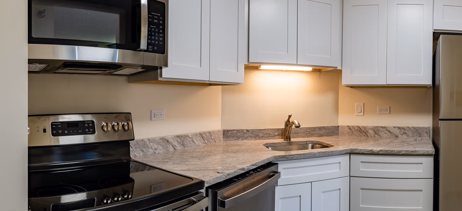 Modern kitchen area with white cabinets, a stainless steel microwave, stove, dishwasher, and refrigerator. The countertop is made of a light-colored stone with a built-in sink and a faucet. Under-cabinet lighting illuminates the countertop.