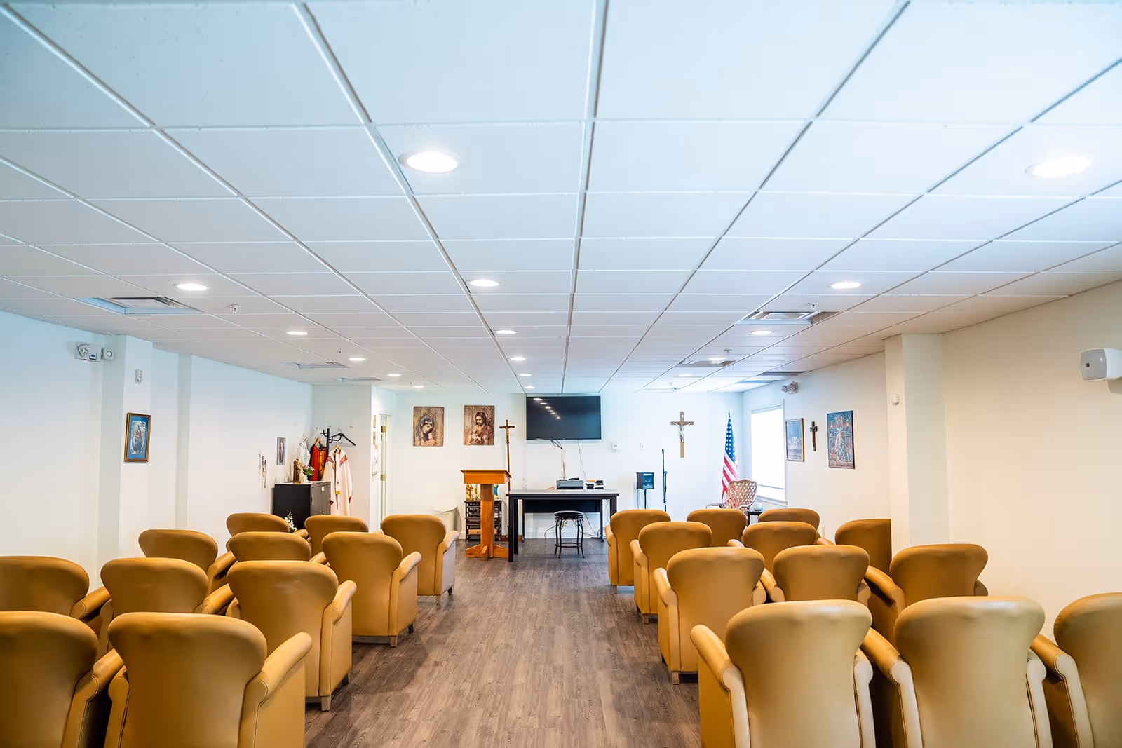 A small chapel or meeting room with rows of tan upholstered chairs facing a podium and a table with a TV mounted on the wall behind it. Religious artwork and crosses are displayed on the walls, and an American flag is positioned near a window on the right side.