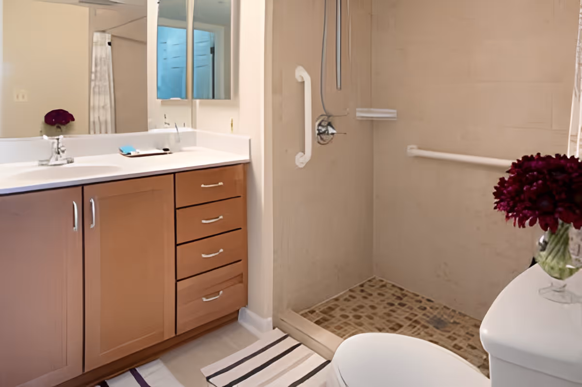A bathroom with a wooden vanity featuring a white countertop and a sink, a large mirror above the sink, a walk-in shower with grab bars and a tiled floor, a toilet, and a vase with dark red flowers on the toilet tank.