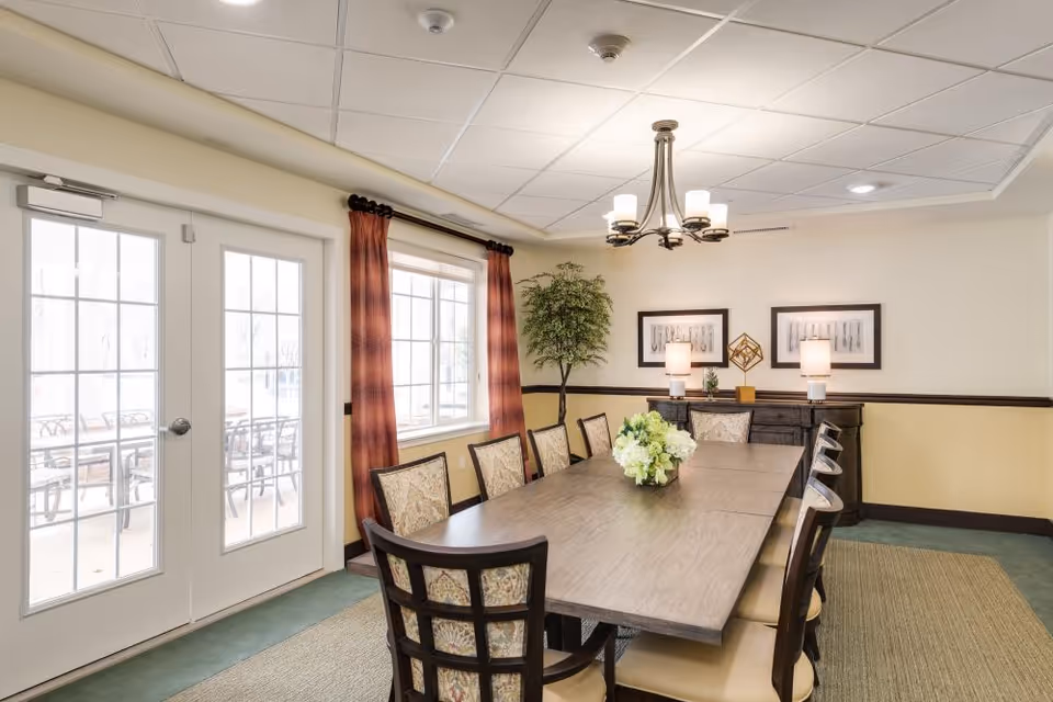 A well-lit dining room with a long rectangular wooden table surrounded by eight cushioned chairs. The room features a large window with red curtains and a glass door leading outside. On the far wall, there is a sideboard with two lamps, decorative items, and framed artwork above it. A chandelier with five lights hangs from the ceiling, and a potted plant is placed in the corner.