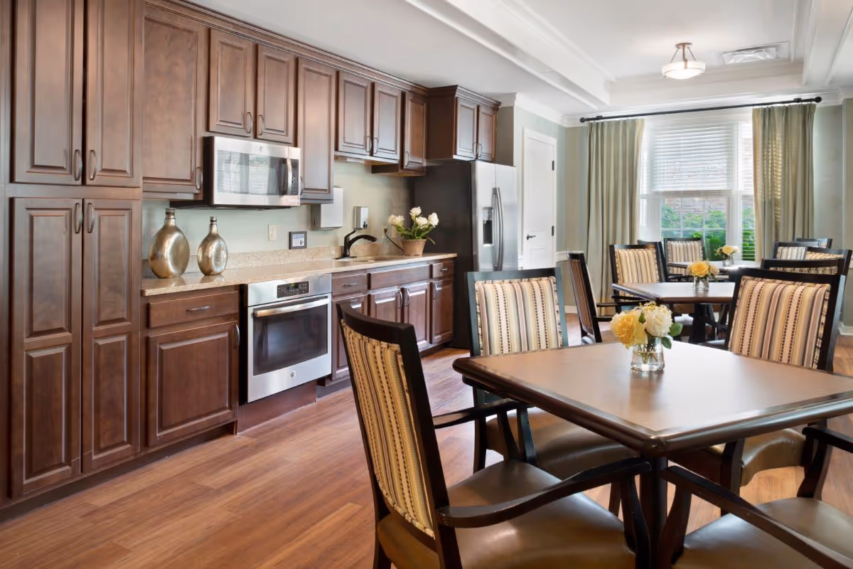 Bright communal dining area with multiple tables and chairs adjacent to a full kitchen with dark wood cabinets and stainless appliances.