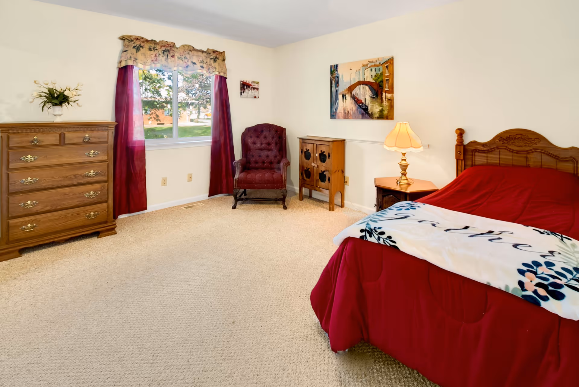 A cozy bedroom with a wooden bed covered in a red comforter and a white blanket with floral designs. There is a wooden nightstand with a lamp beside the bed, a small wooden cabinet, a burgundy upholstered armchair, and a wooden dresser with brass handles. A window with floral valance and burgundy curtains lets in natural light, and a painting of a canal scene hangs on the wall.