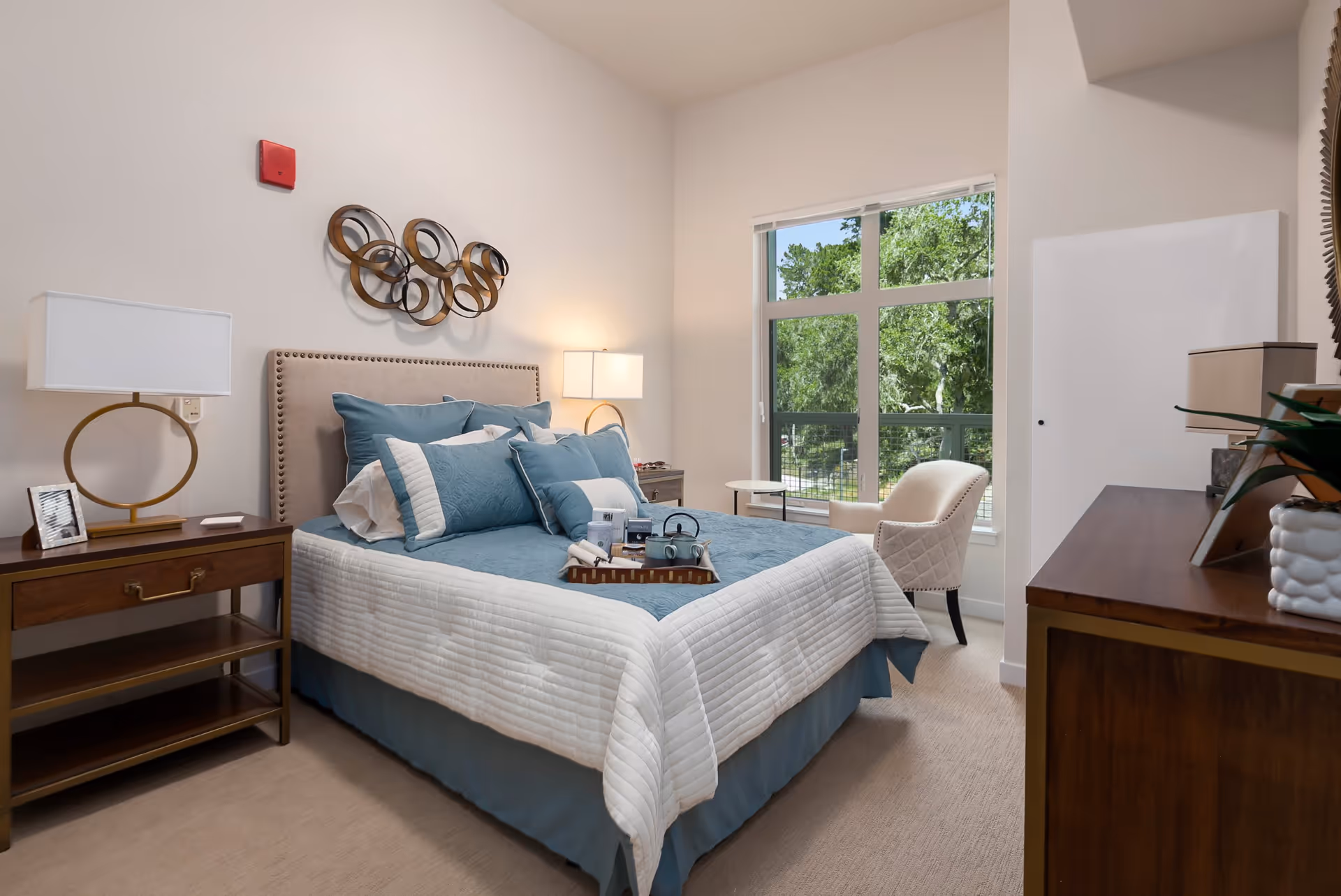 A neatly made bed with blue and white bedding in a bright bedroom. The room features a wooden nightstand with a lamp and a framed photo, a decorative metal wall art above the bed, a window showing green trees outside, a white cushioned chair, and a wooden dresser with a lamp and plant on top.