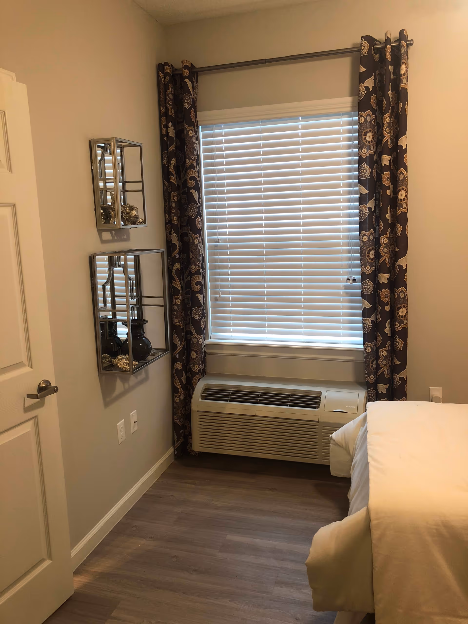 A corner of a bedroom with a window covered by white blinds and dark patterned curtains. Below the window is a wall-mounted air conditioning unit. To the left, there are two decorative mirrored shelves mounted on the wall, holding small decorative items. Part of a bed with white bedding is visible on the right side of the image. The floor is wood, and the walls are painted a light beige color.