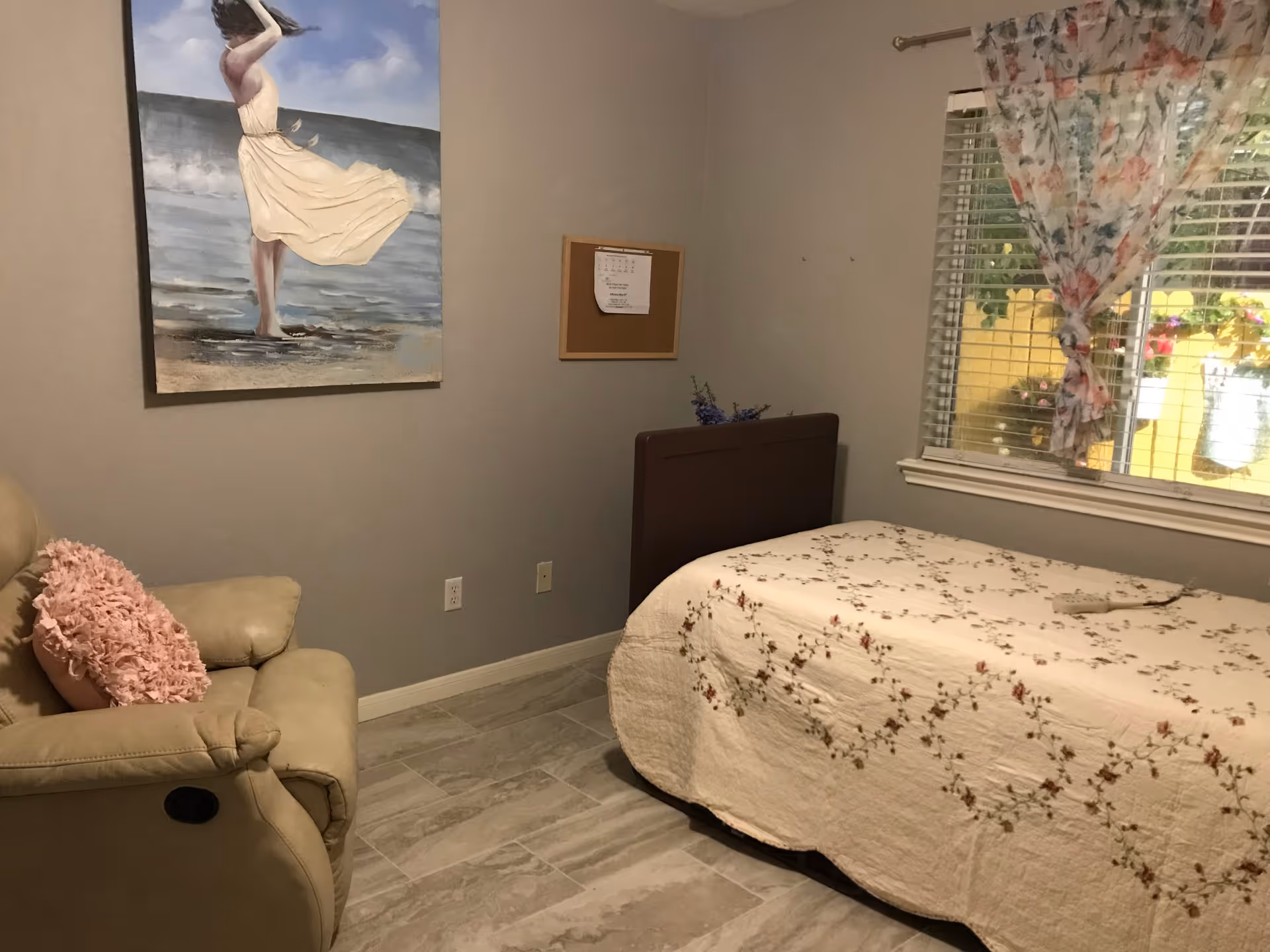 A cozy bedroom with a single bed covered in a floral quilt, a beige recliner chair with a pink textured pillow, a window with floral curtains, a painting of a woman in a flowing dress by the sea, and a small corkboard on the wall.