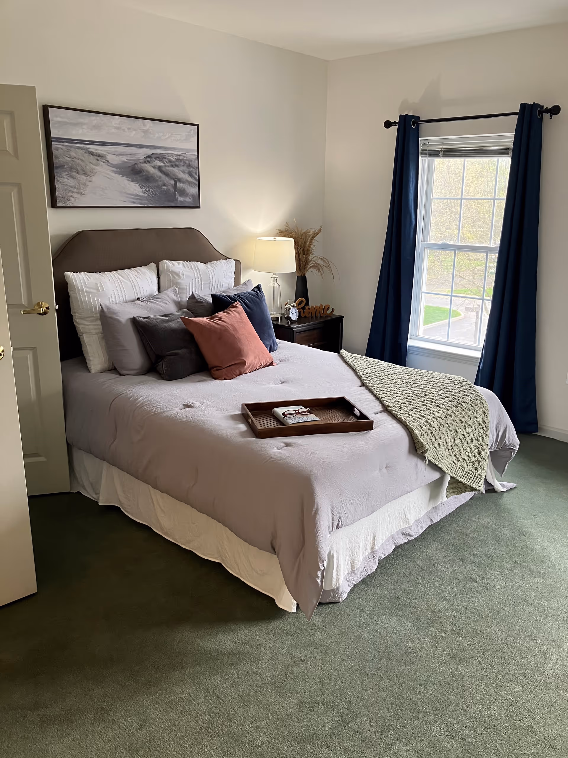 A neatly made bed with multiple pillows in shades of white, gray, navy blue, and pink in a bedroom. A wooden tray with a book and glasses rests on the bed. There is a nightstand with a lamp, a small clock, and decorative items next to the bed. A window with navy blue curtains lets in natural light, and a framed picture of a beach scene hangs on the wall above the bed.