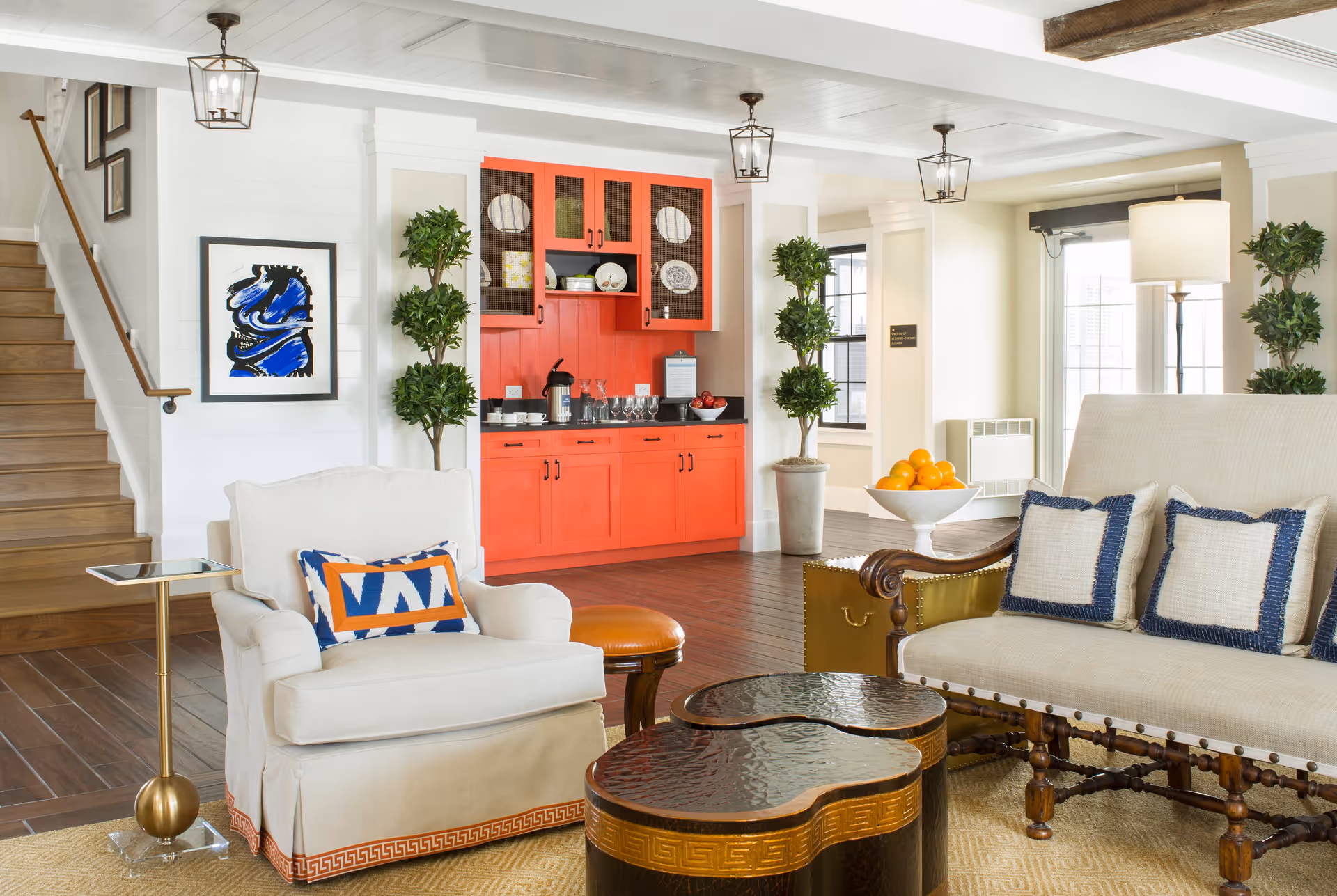 A bright and inviting living room area featuring a white armchair with a colorful cushion, a beige sofa with three blue-bordered pillows, and two uniquely shaped wooden coffee tables. In the background, there is a vibrant red cabinet with glass doors displaying plates and glasses, flanked by two tall potted plants. To the left, wooden stairs lead upward, and the room is well-lit with natural light from windows and ceiling lantern-style lights.