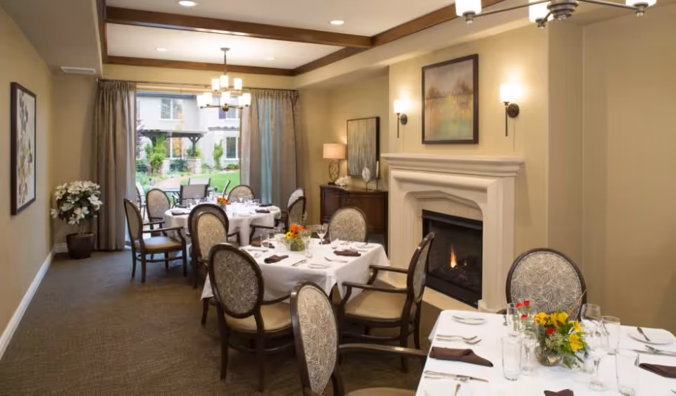 Dining room with tables set for a meal, upholstered chairs, a fireplace, and a view into a courtyard.