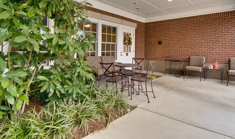 Covered outdoor patio area with brick walls, a white door with a wreath, several cushioned chairs, metal tables and chairs, and green plants along the edge.
