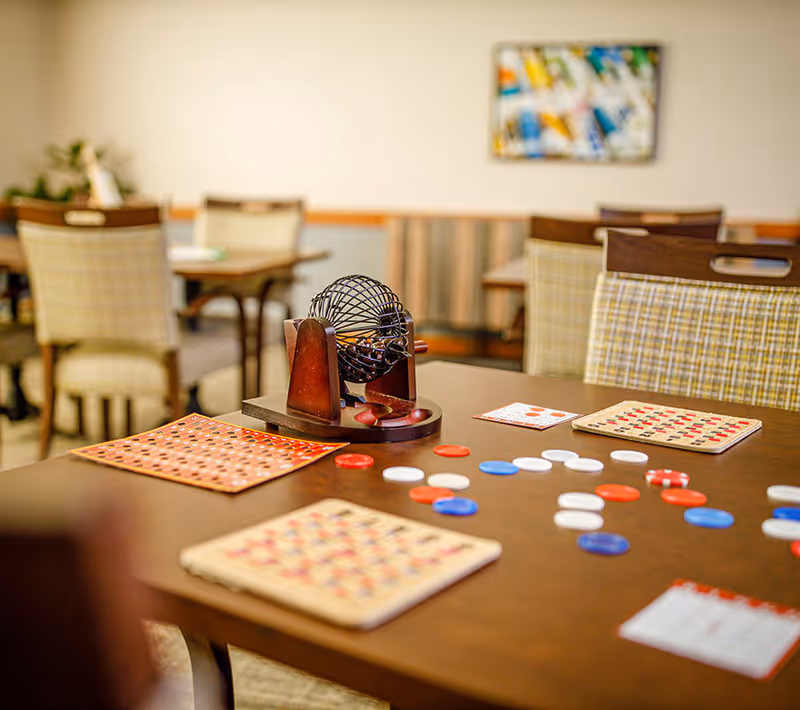 A table in a common area with a bingo cage, bingo cards, and red, white, and blue bingo chips scattered on the table. In the background, there are several chairs and tables with a colorful abstract painting on the wall.