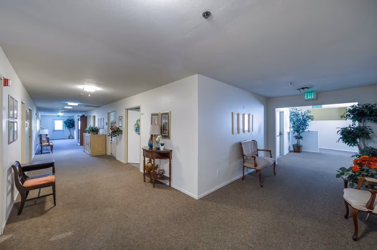 Interior hallway of a senior living facility with beige carpet and white walls. The hallway is decorated with framed pictures, potted plants, and wooden chairs with cushions. There are multiple doors along the hallway and exit signs visible at the far end.