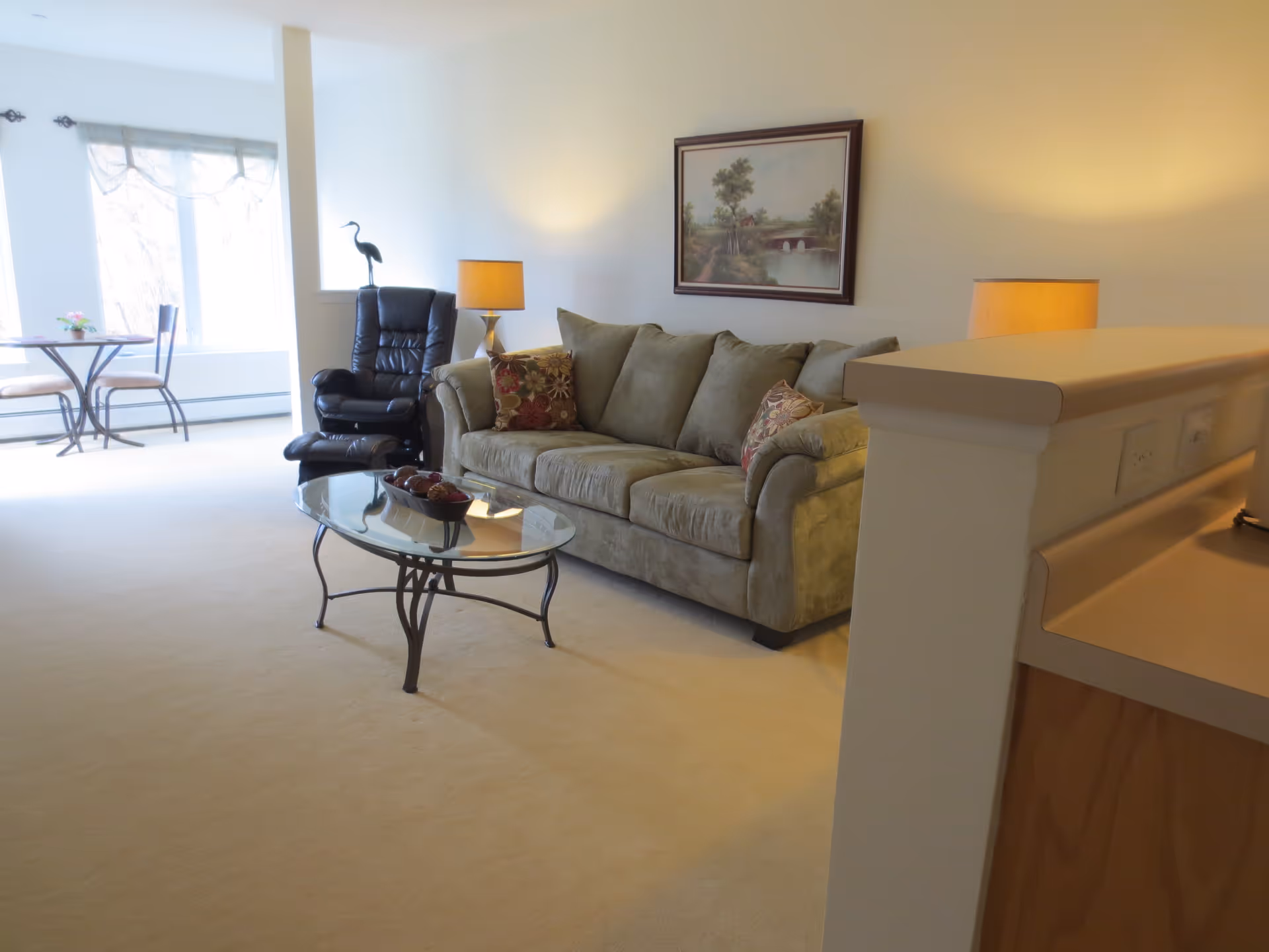 Well-lit living room with a green sofa, glass coffee table, black recliner, table lamps, and a small dining nook by a window.