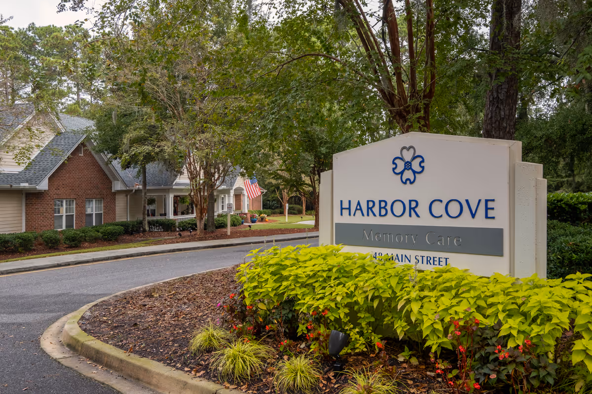 Entrance sign reading 'Harbor Cove Memory Care' by a landscaped driveway with the facility buildings and an American flag in the background.