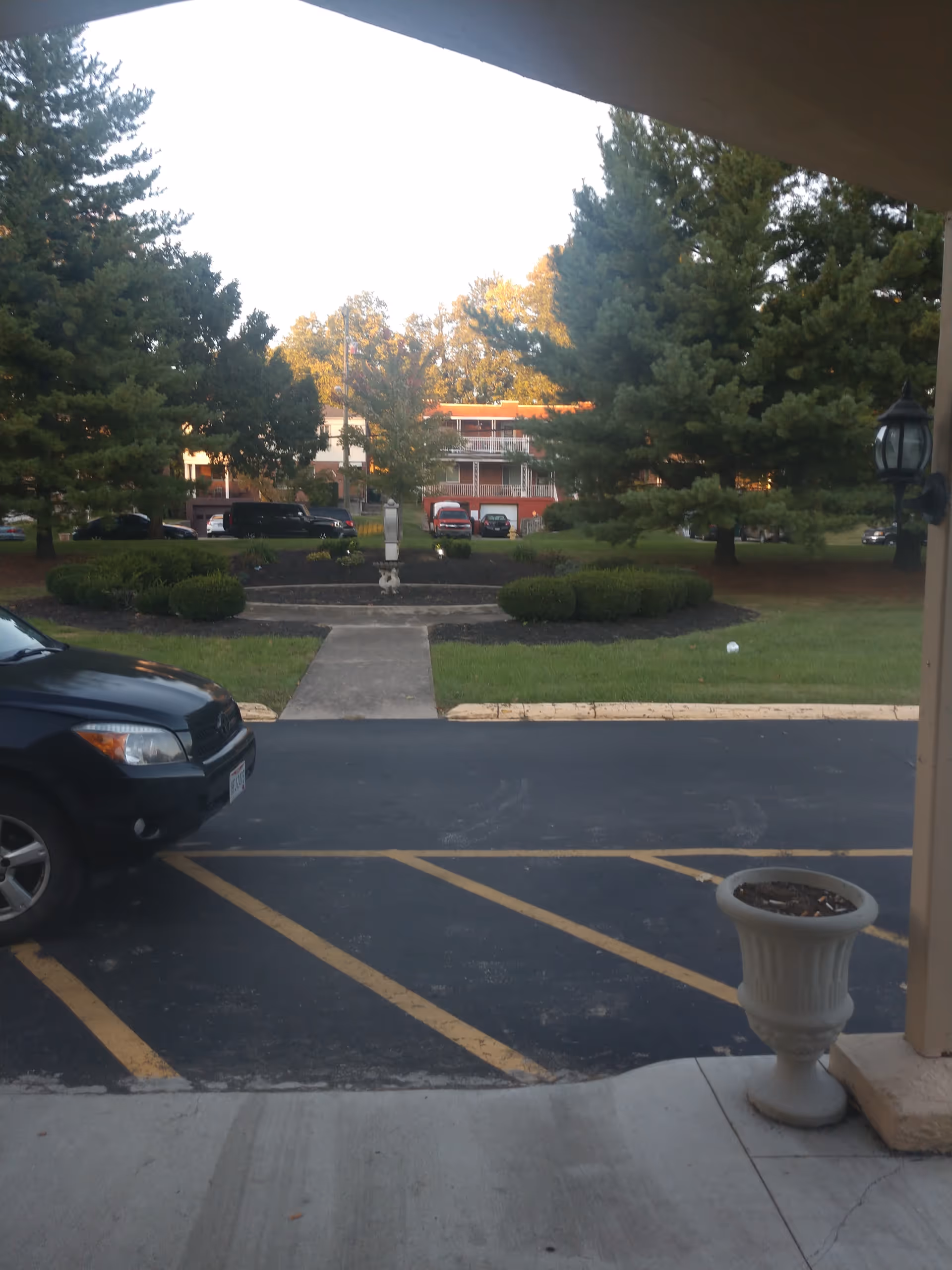 View from a covered entrance across a parking area to a landscaped courtyard with trees, parked cars, and a multi-story building in the background.