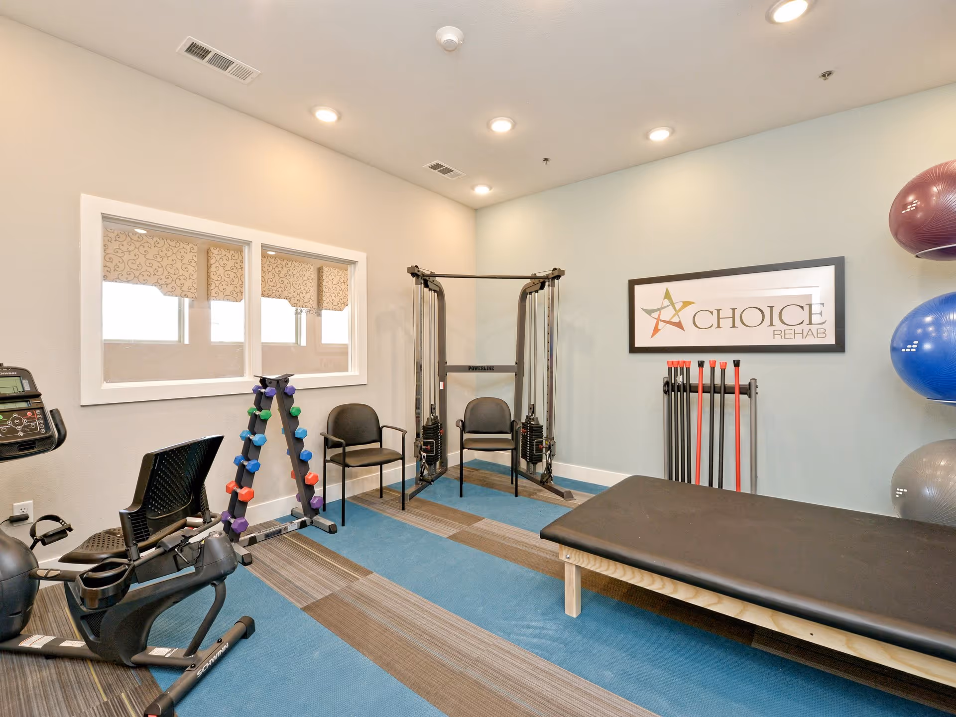 A rehabilitation exercise room with various fitness equipment including a recumbent exercise bike, a rack of colorful dumbbells, a multi-functional cable machine, exercise balls mounted on the wall, and a padded therapy table. Two chairs are placed near the equipment, and a framed sign on the wall reads 'Choice Rehab'. The room has a blue and brown striped carpet and recessed ceiling lights.