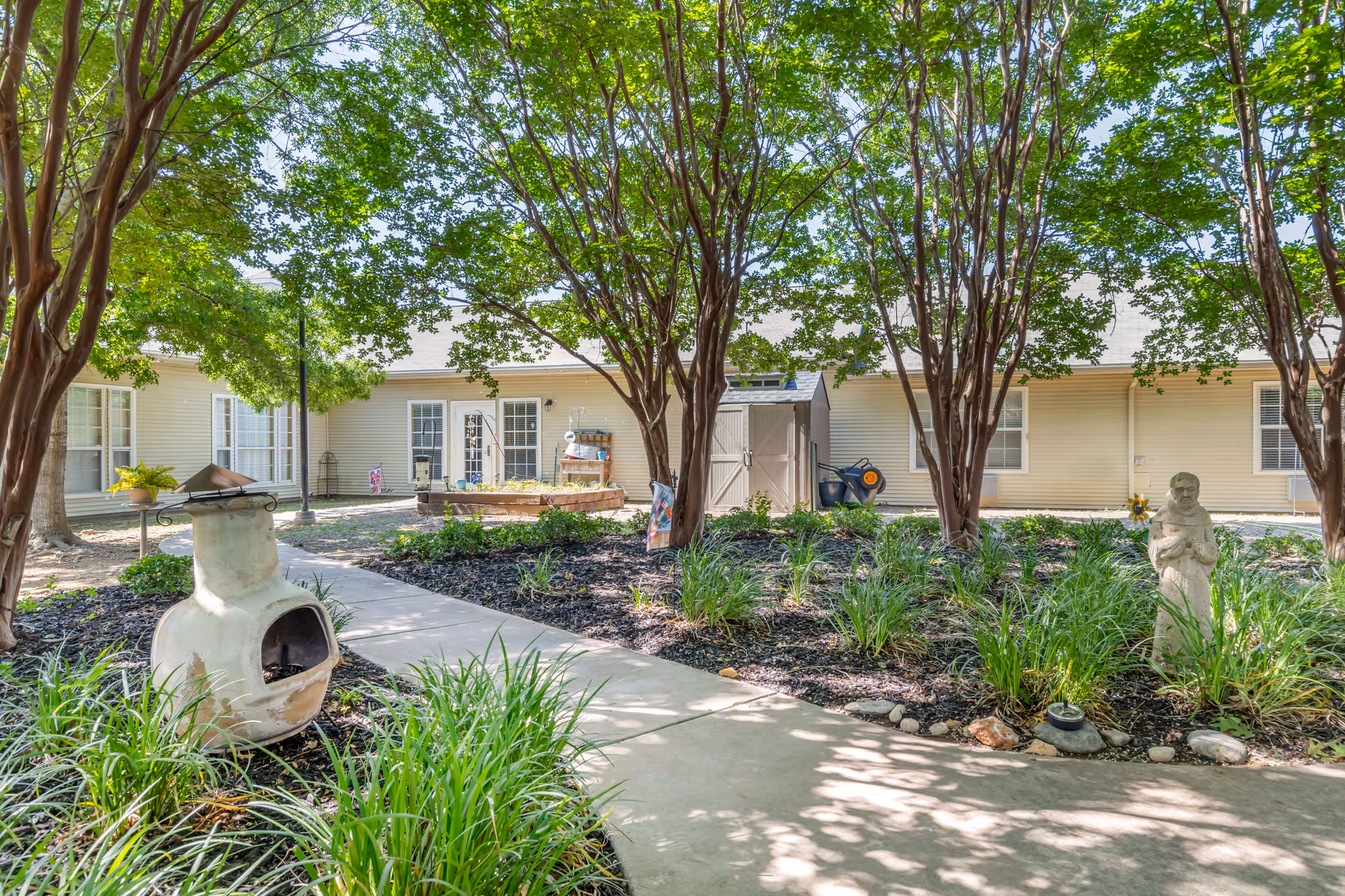 A peaceful outdoor courtyard area with a concrete pathway winding through landscaped garden beds filled with green plants and trees. There is a white chiminea on the left and a small statue on the right. In the background, there is a beige building with multiple windows and doors, a small shed, and some gardening tools.
