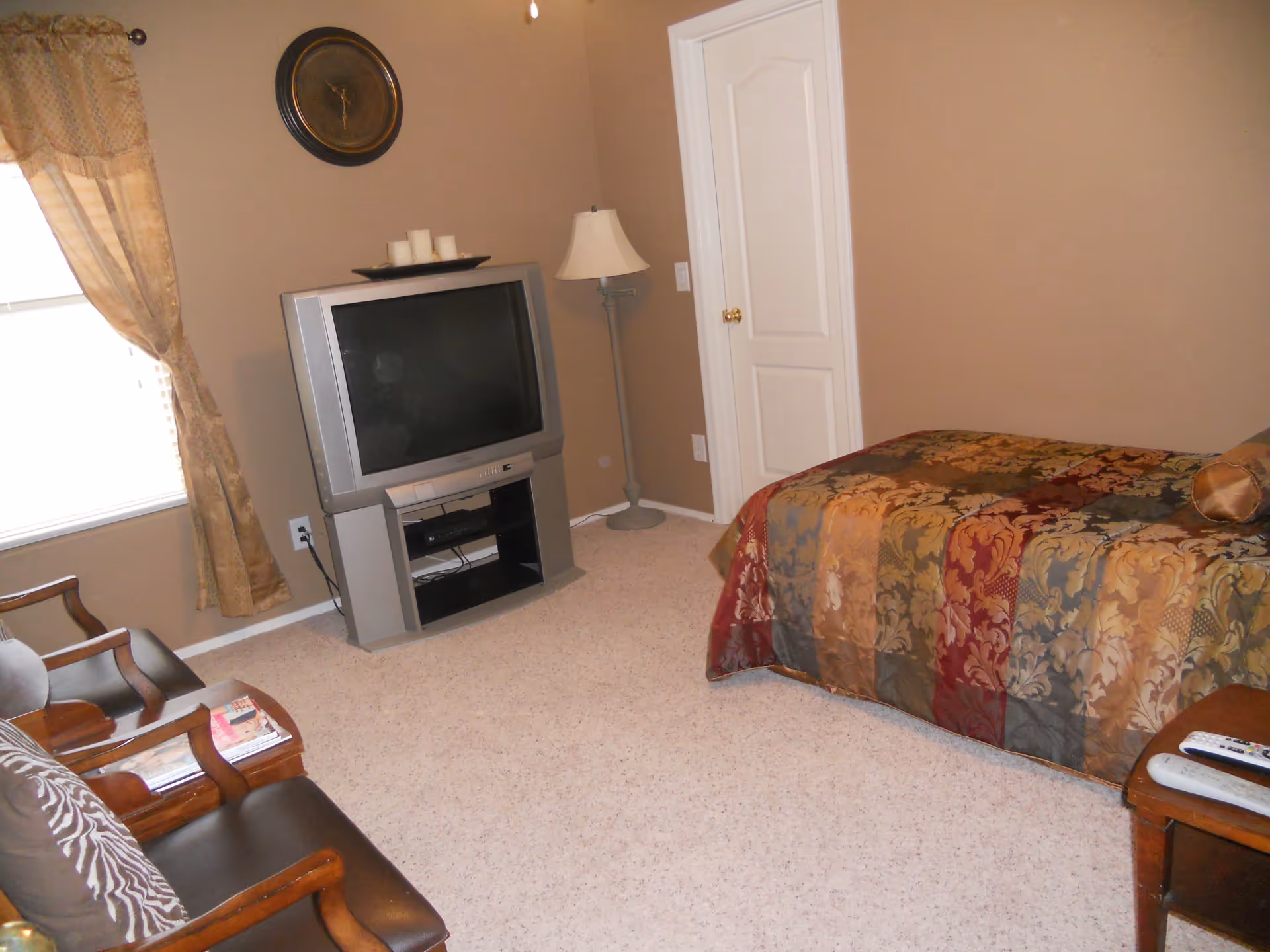A bedroom with beige walls and carpeted floor featuring a bed with a multicolored patterned bedspread, a wooden chair with a cushion and magazines on the armrest, a vintage-style TV on a stand, a floor lamp, a window with gold curtains, and a wall clock above the TV.