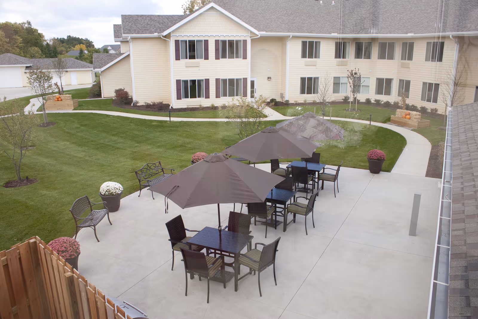 Outdoor patio area at Amber Park Senior Living with several tables and chairs under large umbrellas, surrounded by green grass, potted flowers, benches, and a beige two-story building in the background.