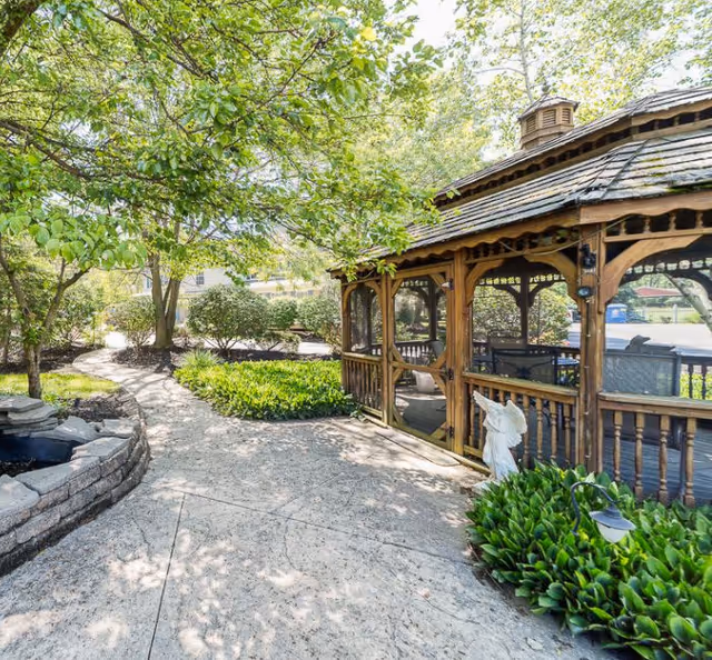 A peaceful outdoor garden area with a stone pathway leading past a small pond and a wooden gazebo surrounded by greenery and trees, with sunlight filtering through the leaves.