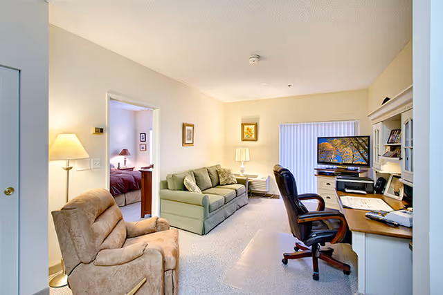 A cozy living area in a senior living facility with a green sofa, a beige recliner, and a wooden desk with a black office chair. A flat-screen TV is on the desk, and there is a lamp and framed artwork on the walls. An open doorway reveals a bedroom with a bed and nightstand lamp.