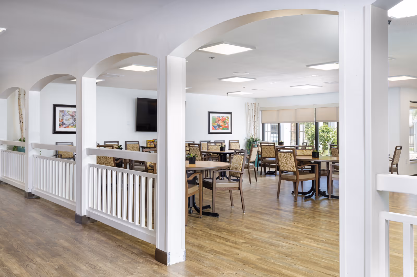 A bright dining room with wooden floors and multiple tables and chairs arranged neatly. The room features white walls, large windows with beige blinds, framed artwork on the walls, and a flat-screen TV mounted on one wall. White columns and arches partially separate the dining area from the adjacent space.