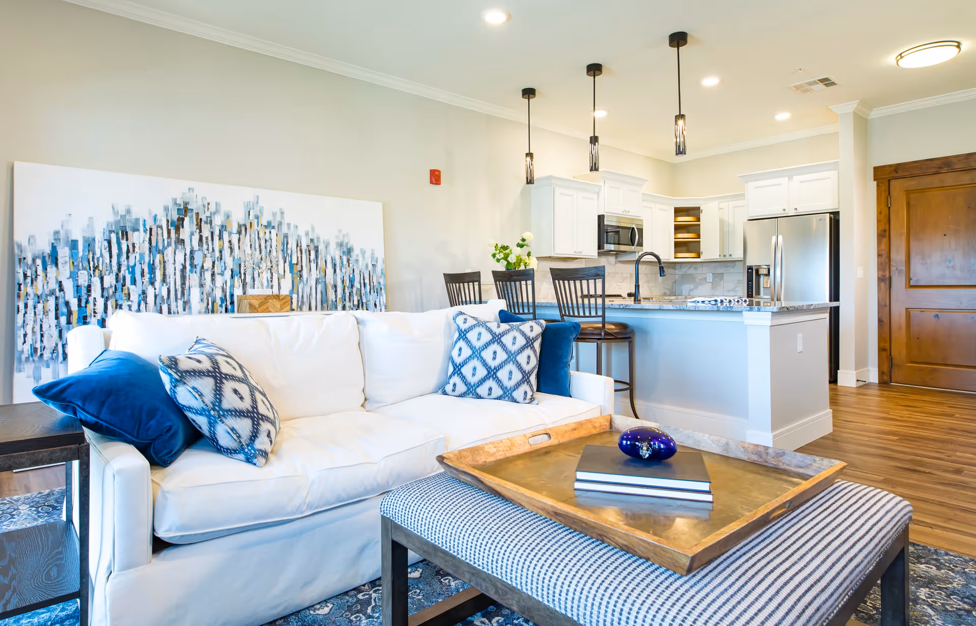 Bright and modern living room with a white sofa adorned with blue and patterned pillows, a wooden coffee table with books and a decorative item, and a kitchen area in the background featuring white cabinets, a stainless steel refrigerator, a microwave, and a kitchen island with bar stools and pendant lights.