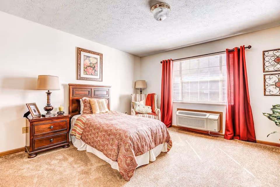 A cozy bedroom with a single bed covered in a patterned bedspread and multiple pillows. Next to the bed is a wooden nightstand with a lamp, a framed photo, and a candle. A cushioned armchair with a pillow and a red throw blanket is positioned near a large window with red curtains. The room has beige carpet and light-colored walls, with a framed floral picture hanging above the bed.