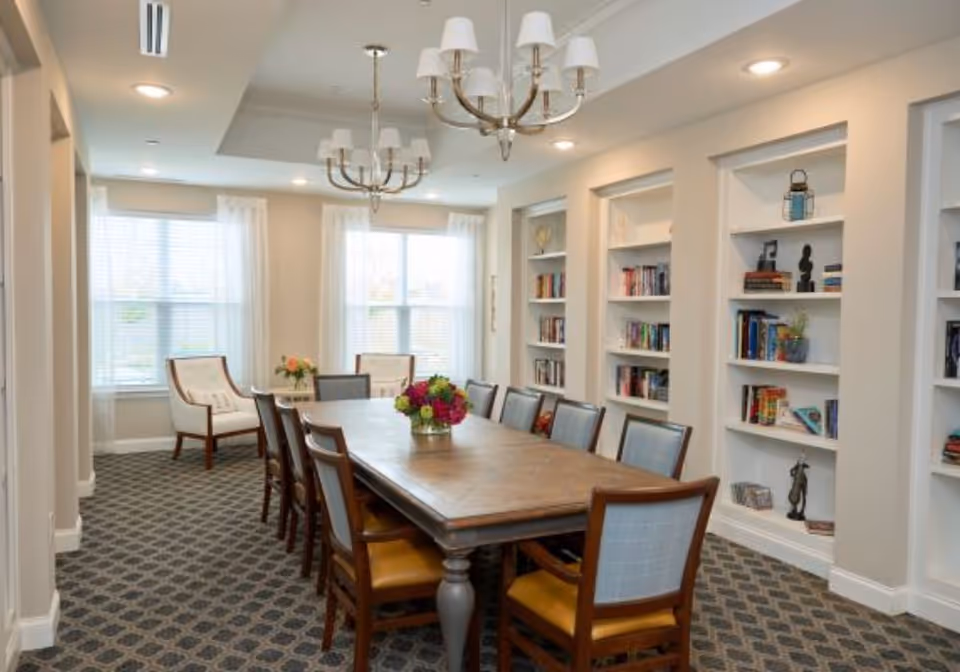 Bright dining room with a long wooden table surrounded by chairs, chandeliers overhead, and built-in bookshelves along one wall.