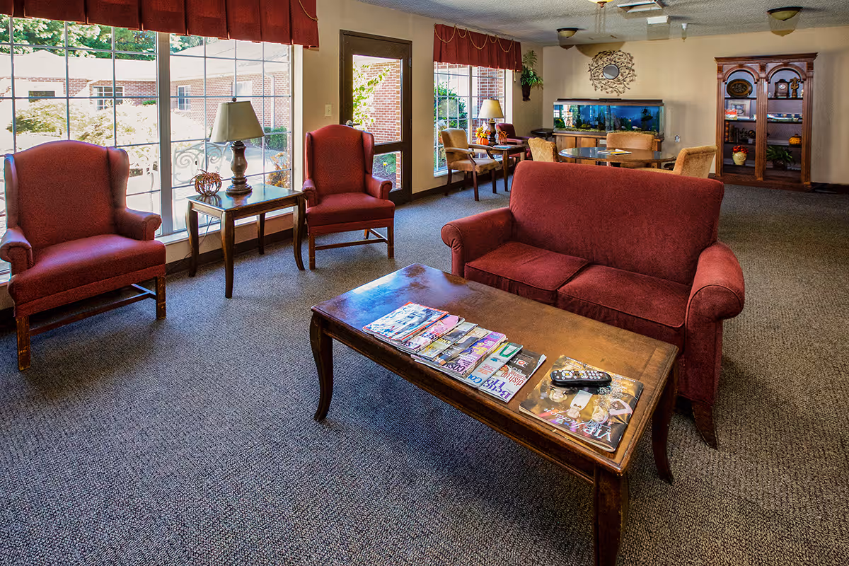 A cozy living room area in a senior living facility with red upholstered chairs and a sofa, a wooden coffee table with magazines and remote controls, large windows with red valances letting in natural light, a wooden cabinet with decorative items, and a fish tank in the background.