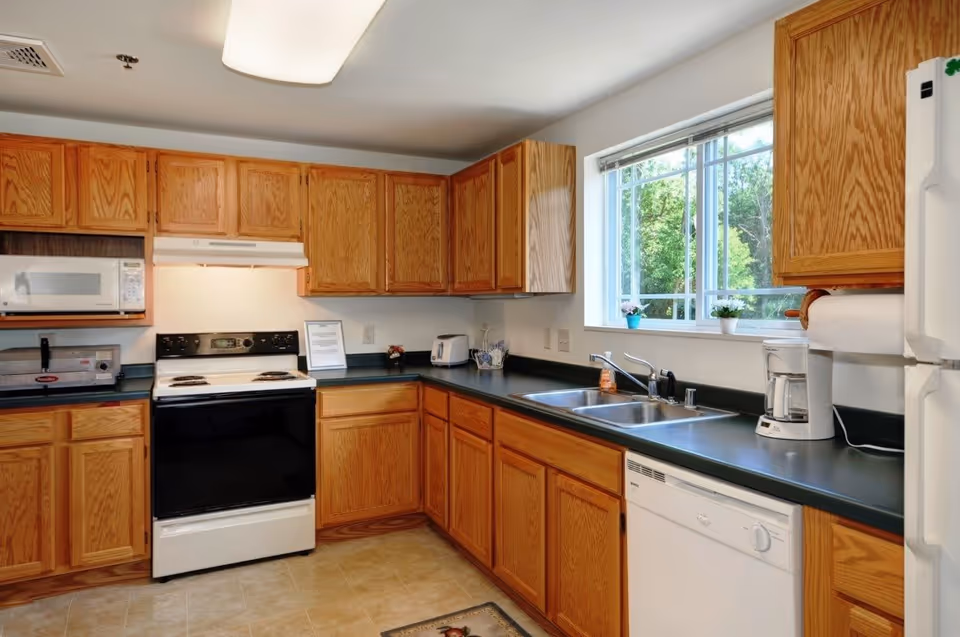 A kitchen with wooden cabinets, a white stove with a black oven door, a microwave, a toaster, a coffee maker, a double sink under a window showing green trees outside, and a white dishwasher. The countertops are dark green and the floor is tiled.